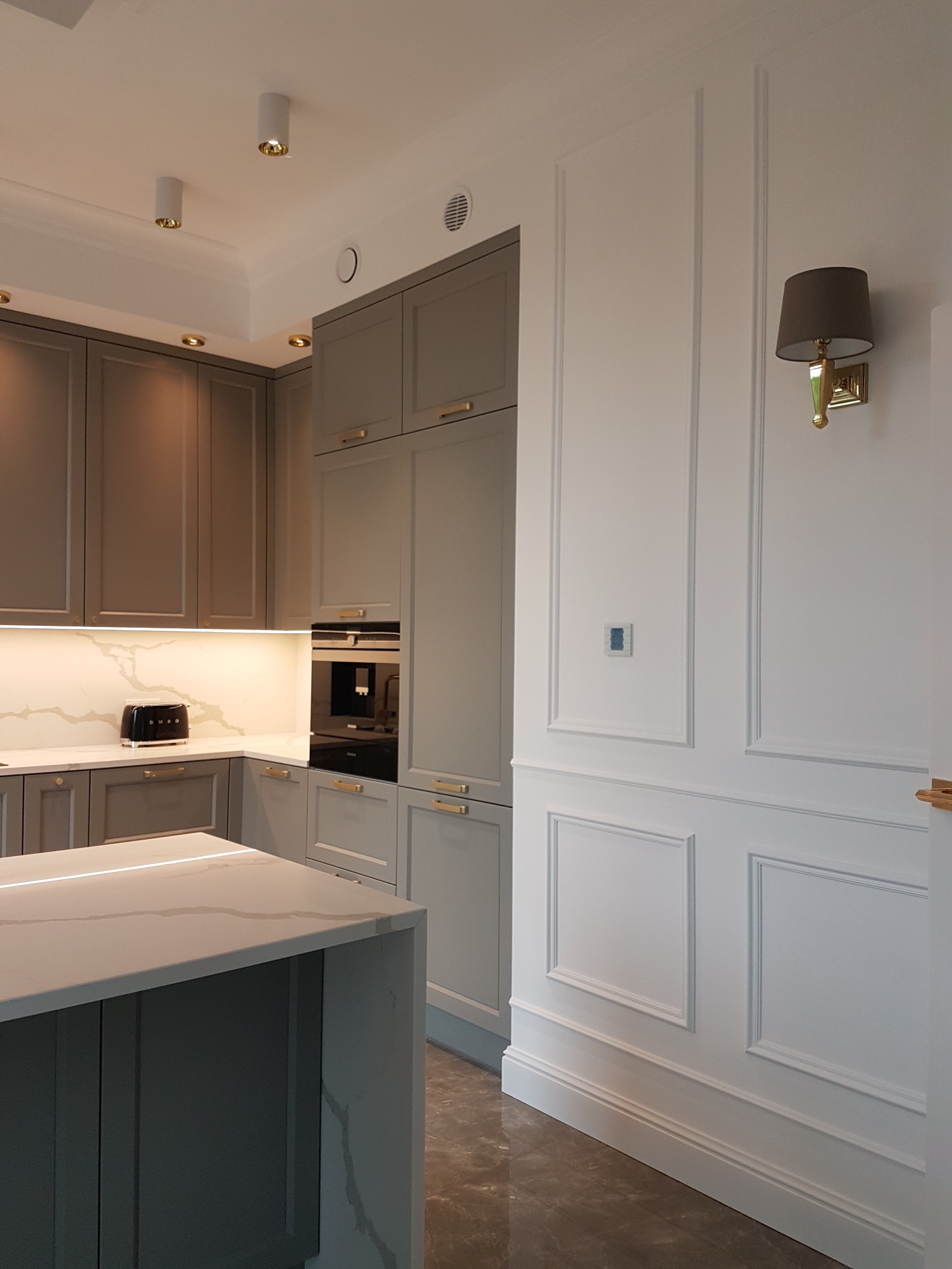 Nowoczesna kuchnia w odcieniach szarości z wyspą i białymi ścianami z dekoracyjnymi panelami, oświetlona punktowo i kinkietem.