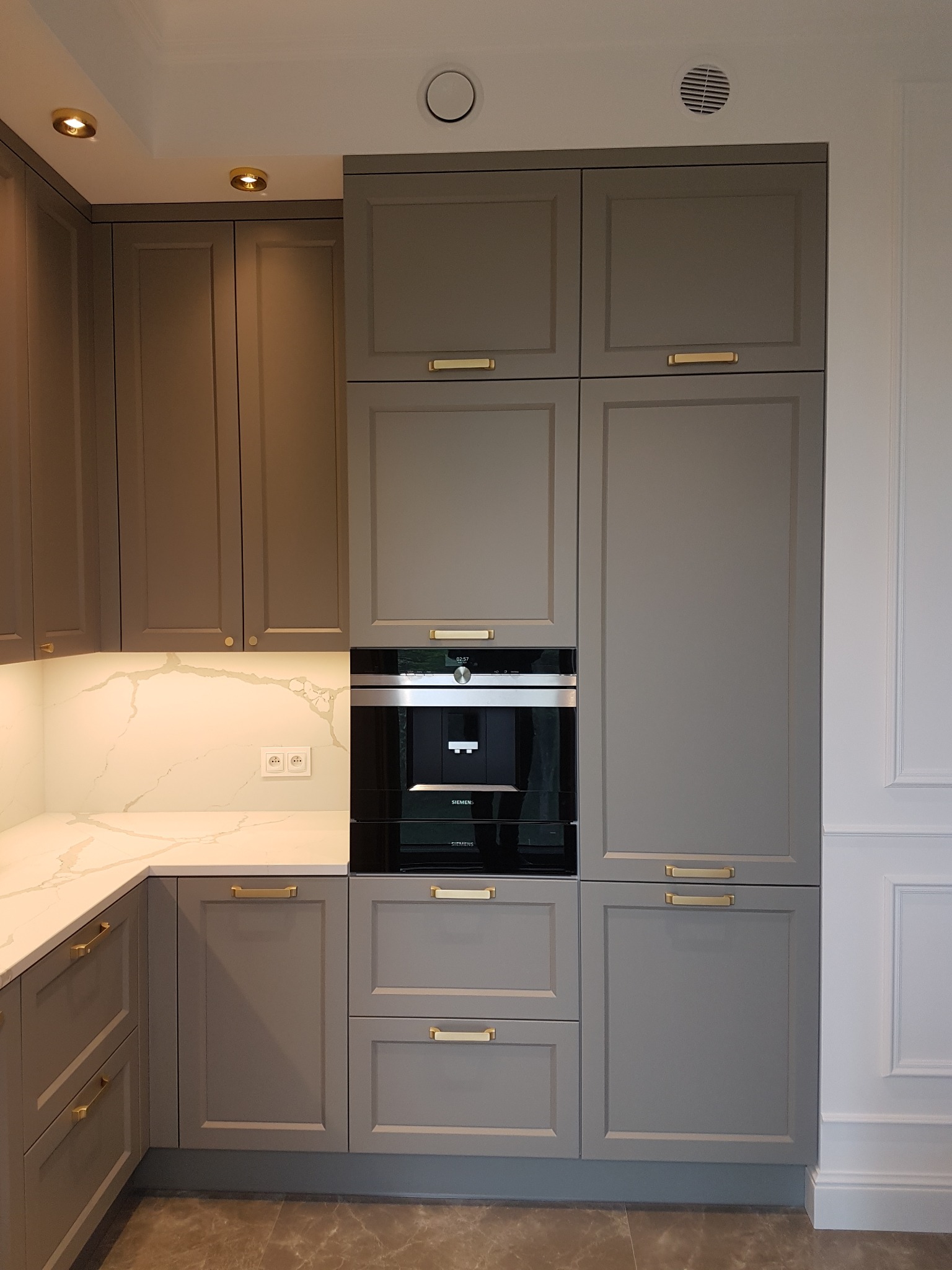 Szare, klasyczne szafki kuchenne z złotymi uchwytami, marmurowy blat z podświetleniem LED, zabudowany ekspres do kawy Siemens