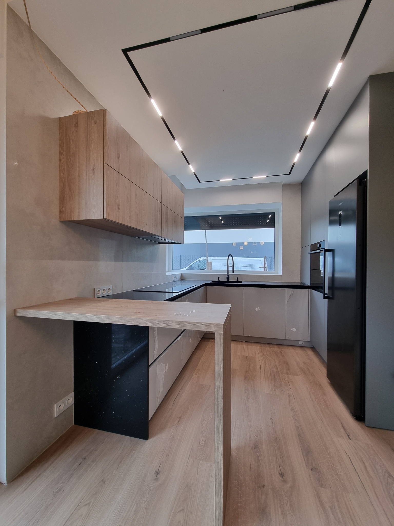 Nowoczesna kuchnia z drewnianymi szafkami wiszącymi, czarnym blatem, wyspą z drewnianym blatem i czarnym panelem dekoracyjnym, oświetleniem LED w suficie i drewnianą podłogą.