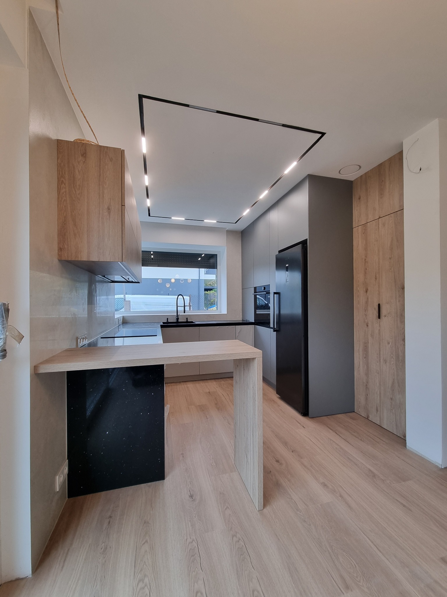 Nowoczesna kuchnia z wyspą, szare szafki z czarną lodówką, drewniane akcenty i minimalistyczne oświetlenie LED.