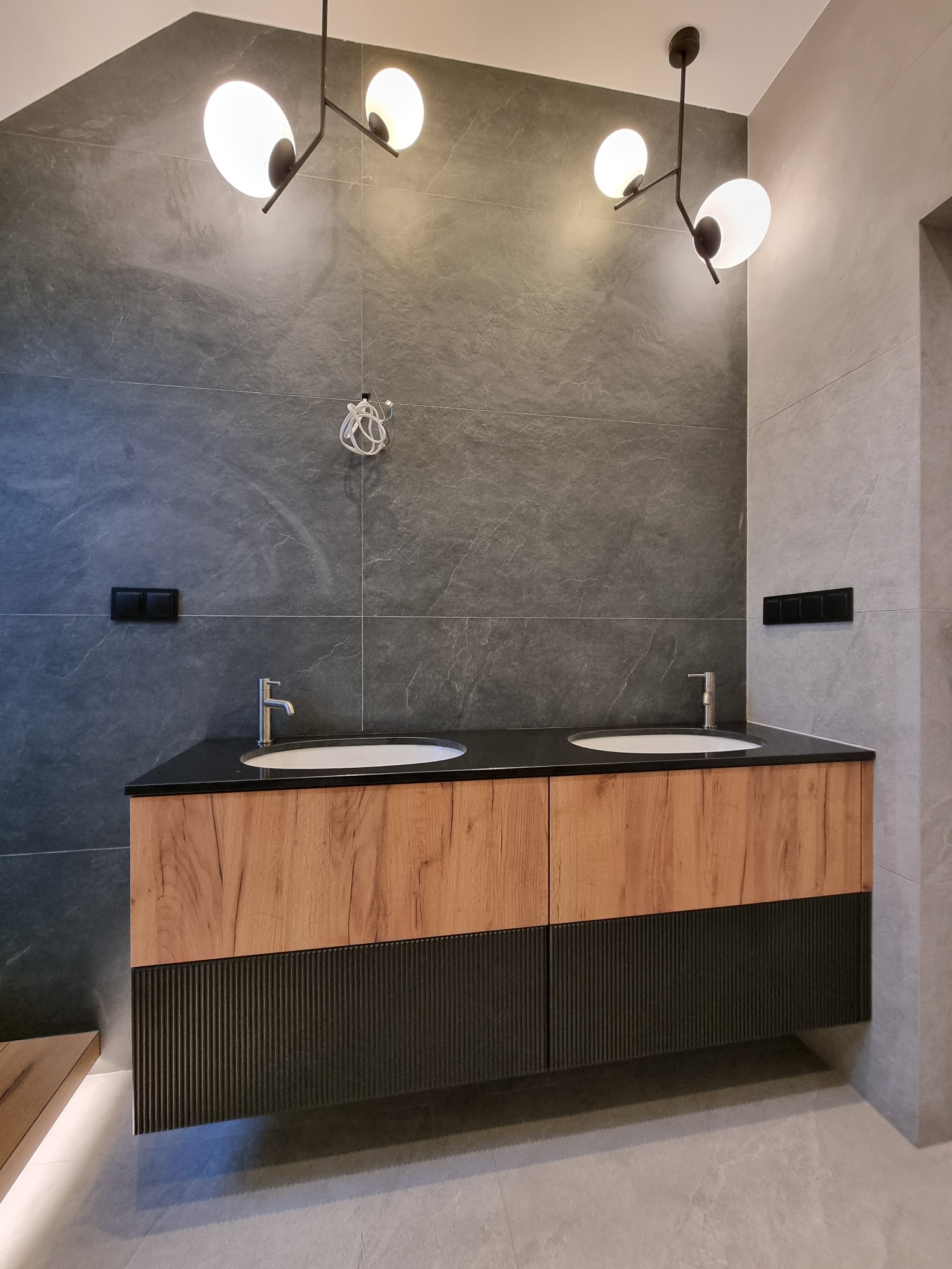 Nowoczesna łazienka z podwójną umywalką na szafce z drewnopodobnym frontem i czarnym, żłobionym dołem, oświetlona lampami sufitowymi z kulistymi kloszami, na ścianach szare płytki imitujące kamień,...