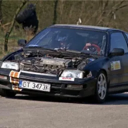 Czarny samochód wyścigowy Honda CRX z otwartą maską, widoczny silnik i kierowca w kasku na torze wyścigowym, naklejki sponsorskie na karoserii i pomarańczowe pasy na przednim zderzaku.