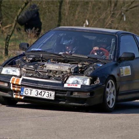 Czarny samochód wyścigowy Honda CRX z otwartą maską, widoczny silnik i kierowca w kasku na torze wyścigowym, naklejki sponsorskie na karoserii i pomarańczowe pasy na przednim zderzaku.