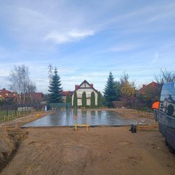 HEXIUM SPÓŁKA Z OGRANICZONĄ ODPOWIEDZIALNOŚCIĄ - Wylana płyta fundamentowa pod dom jednorodzinny, widoczne szalunki, zbrojenie i poziomica laserowa. W tle dom w stylu dworkowym i błękitne niebo.