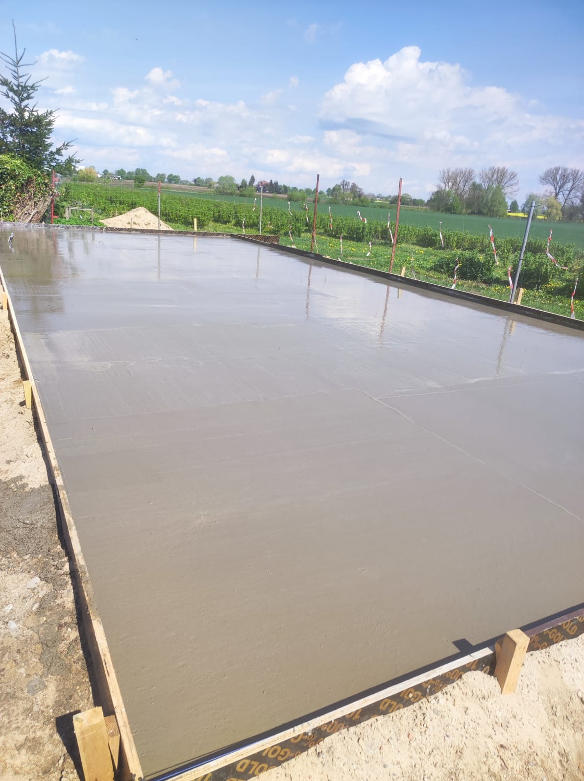 Świeżo wylana, gładka płyta fundamentowa na tle zielonego pola i błękitnego nieba. Widoczne drewniane szalunki i oznaczenia geodezyjne.