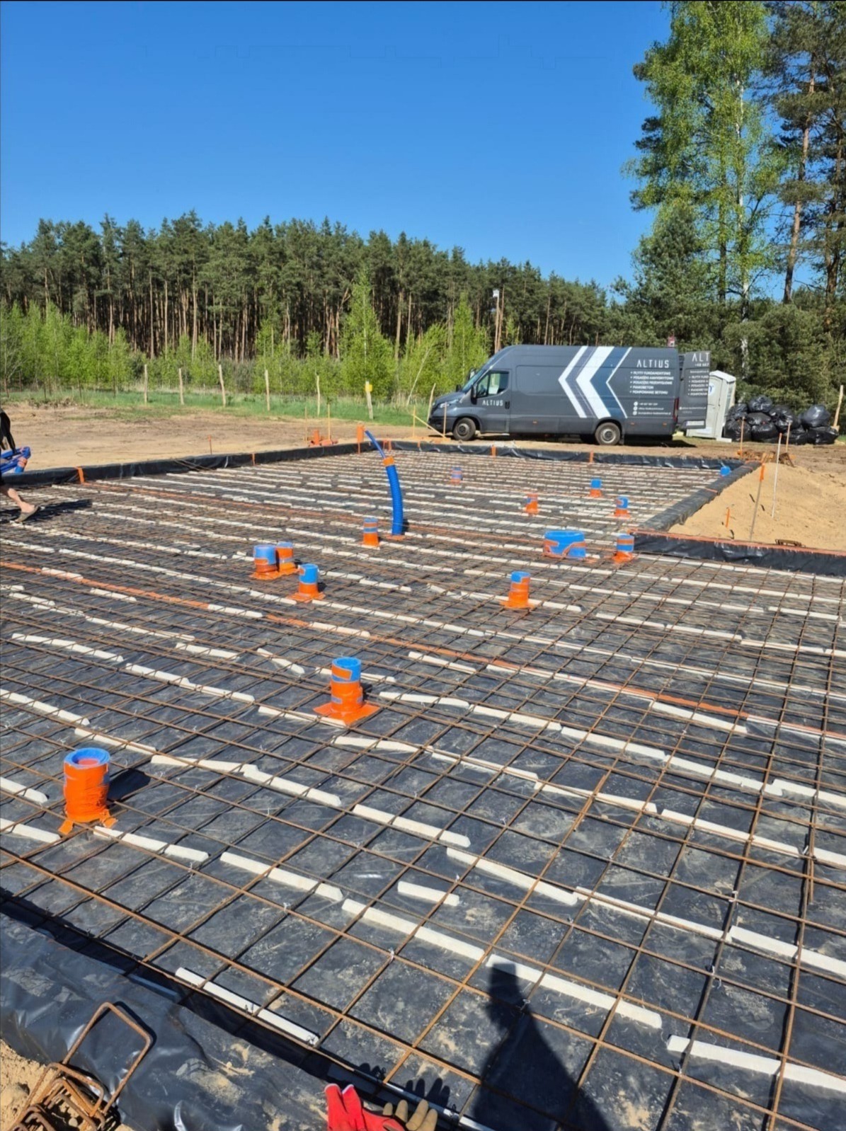 Przygotowanie płyty fundamentowej: stalowe zbrojenie, izolacja termiczna, rury ogrzewania podłogowego. W tle las i samochód firmy Altius.