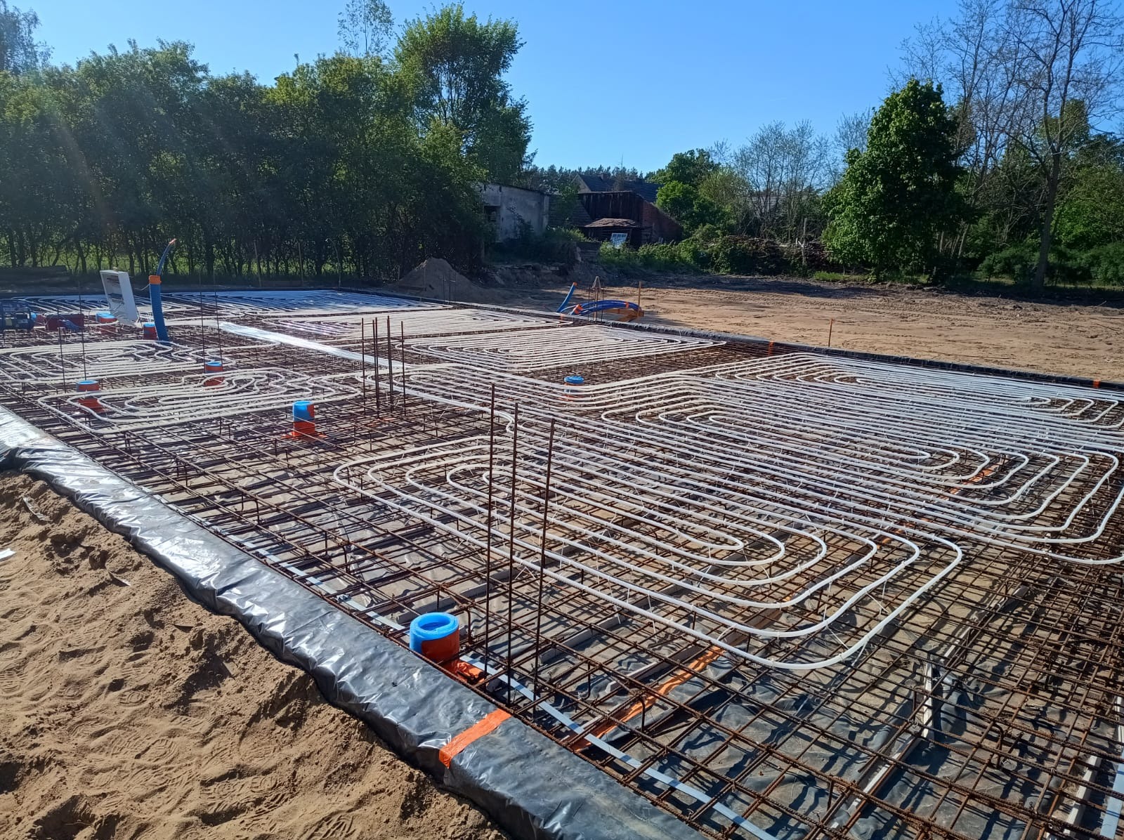 Zbrojenie fundamentów z instalacją ogrzewania podłogowego, widoczne rury w kształcie meandrów, pionowe pręty zbrojeniowe i obrzeża izolacyjne. Dzień, teren budowy.