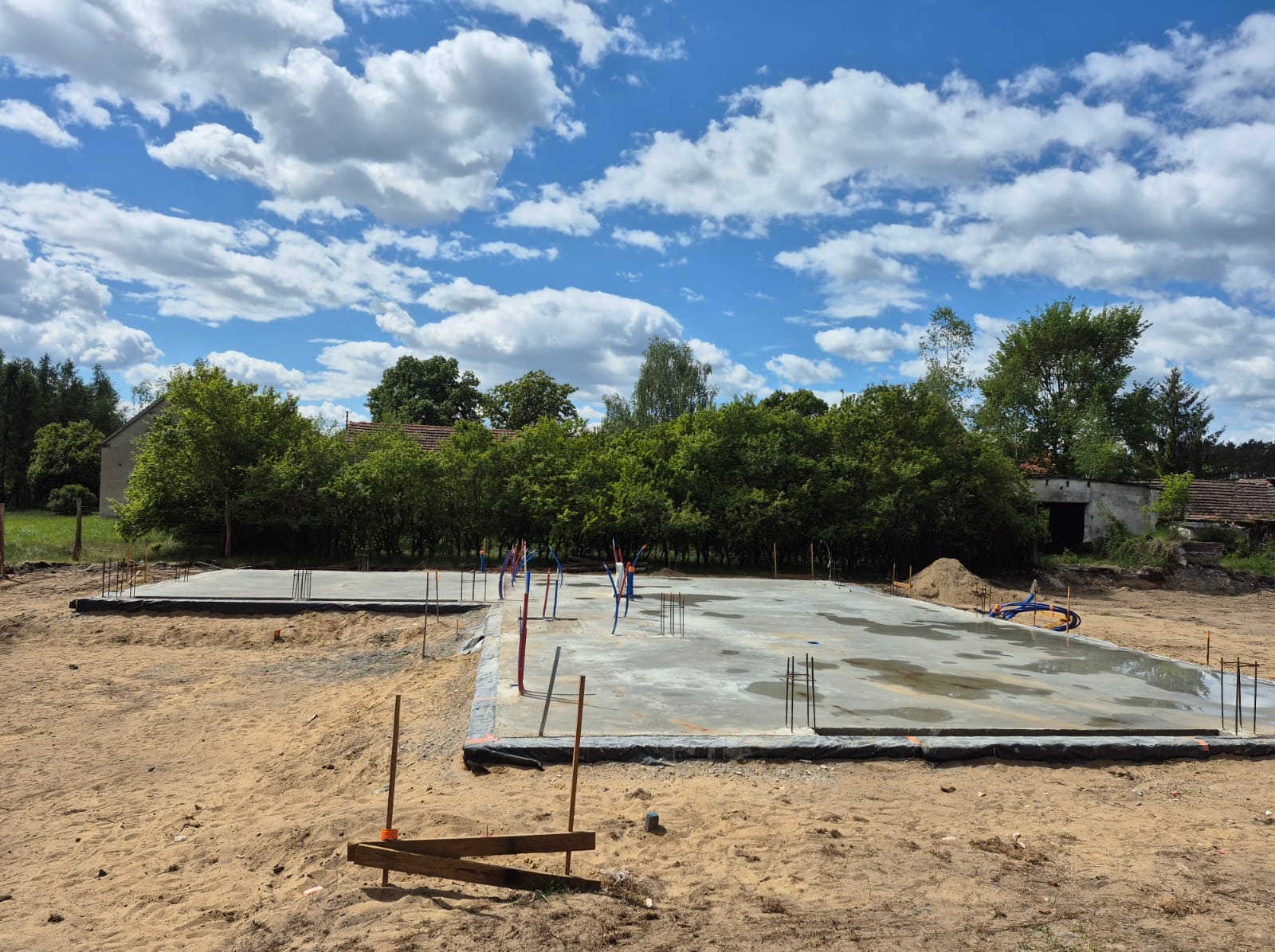 Wylane, betonowe fundamenty domu jednorodzinnego z wystającymi prętami zbrojeniowymi i instalacją wodną, na tle zieleni i błękitnego nieba z chmurami.