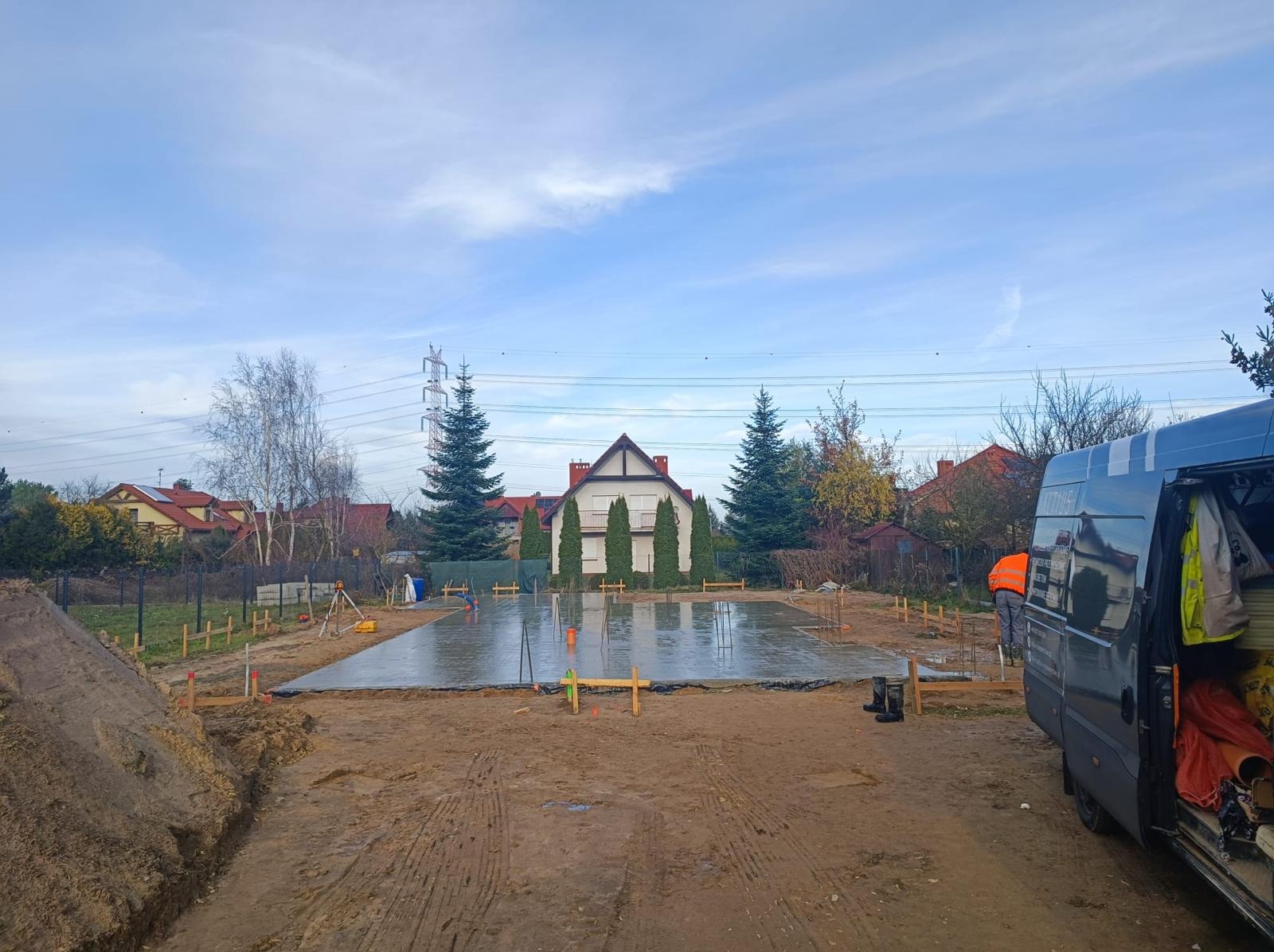 Wylana płyta fundamentowa pod dom jednorodzinny, widoczne szalunki, zbrojenie i poziomica laserowa. W tle dom w stylu dworkowym i błękitne niebo.