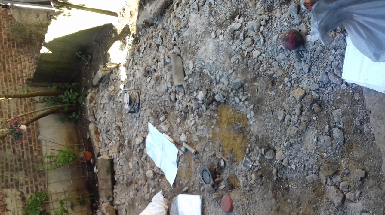 Przygotowanie podłoża pod wylewkę na zewnątrz, widoczne gruz, piasek, rury, kabel na bębnie i częściowo widoczna ściana budynku z roślinami.