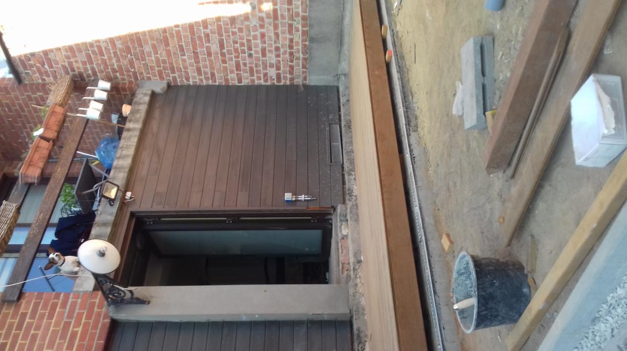 Widok z góry na montaż okna balkonowego z drewnianą podłogą tarasu, widoczne narzędzia i materiały budowlane, ceglana ściana z lampą i pieskiem w tle.