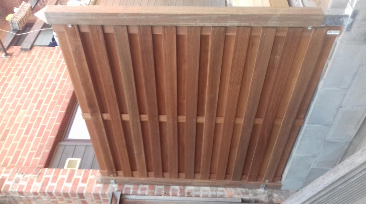 Drewniana osłona balkonu z pionowych desek, widok z dołu, fragment muru z czerwonej cegły w tle.