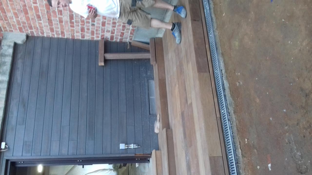 Drewniany taras z odpływem liniowym, przylegający do ściany z okładziną imitującą drewno, częściowo widoczna osoba.