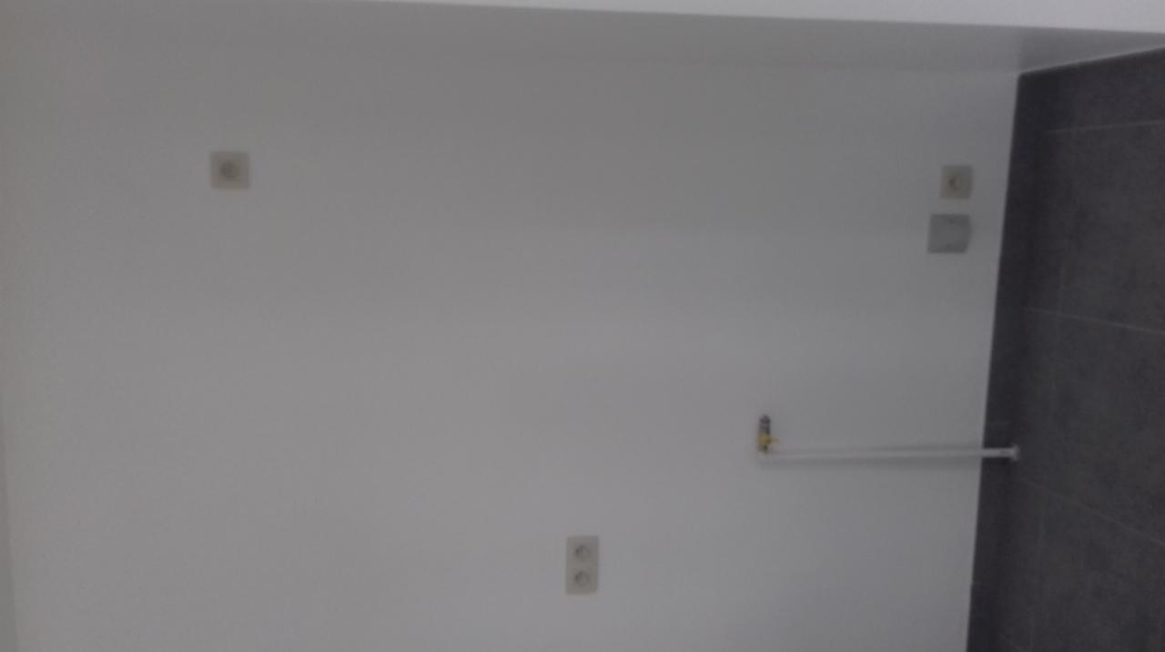 Świeżo wykafelkowana ściana w łazience z widocznymi gniazdkami elektrycznymi, rurą wodną i szarymi płytkami.