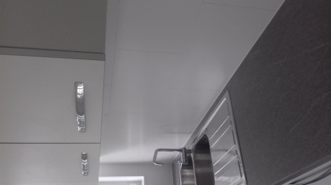 Fragment kuchni z białymi szafkami, chromowanymi uchwytami, stalowym zlewem i ciemnoszarym blatem, widok z dołu.