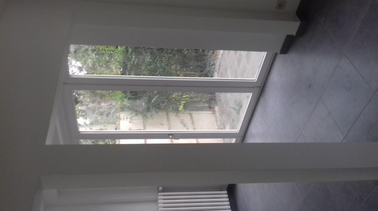 Białe dwuskrzydłowe okno z widokiem na zieleń ogrodu, szare płytki podłogowe i biały grzejnik pod oknem.