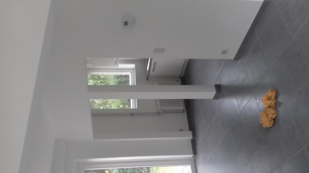 Wnętrze wykończonego pokoju z szarą podłogą, białymi ścianami i widocznymi oknami oraz grzejnikami; na podłodze leży zwinięta ścierka.