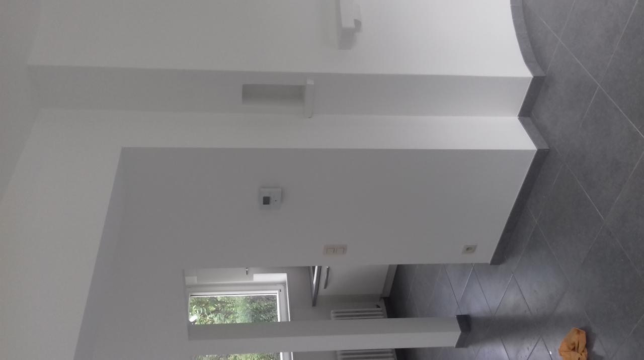 Wnętrze po remoncie: białe ściany z wnękami, szara podłoga z płytek, widoczne okno z zielenią za nim.