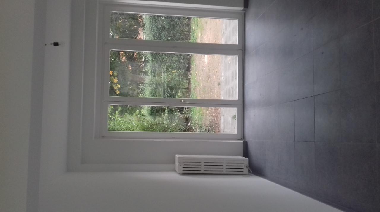 Wnętrze z trzyskrzydłowym oknem PCV, widok na zieleń ogrodu, szare płytki podłogowe i biały grzejnik pod oknem.