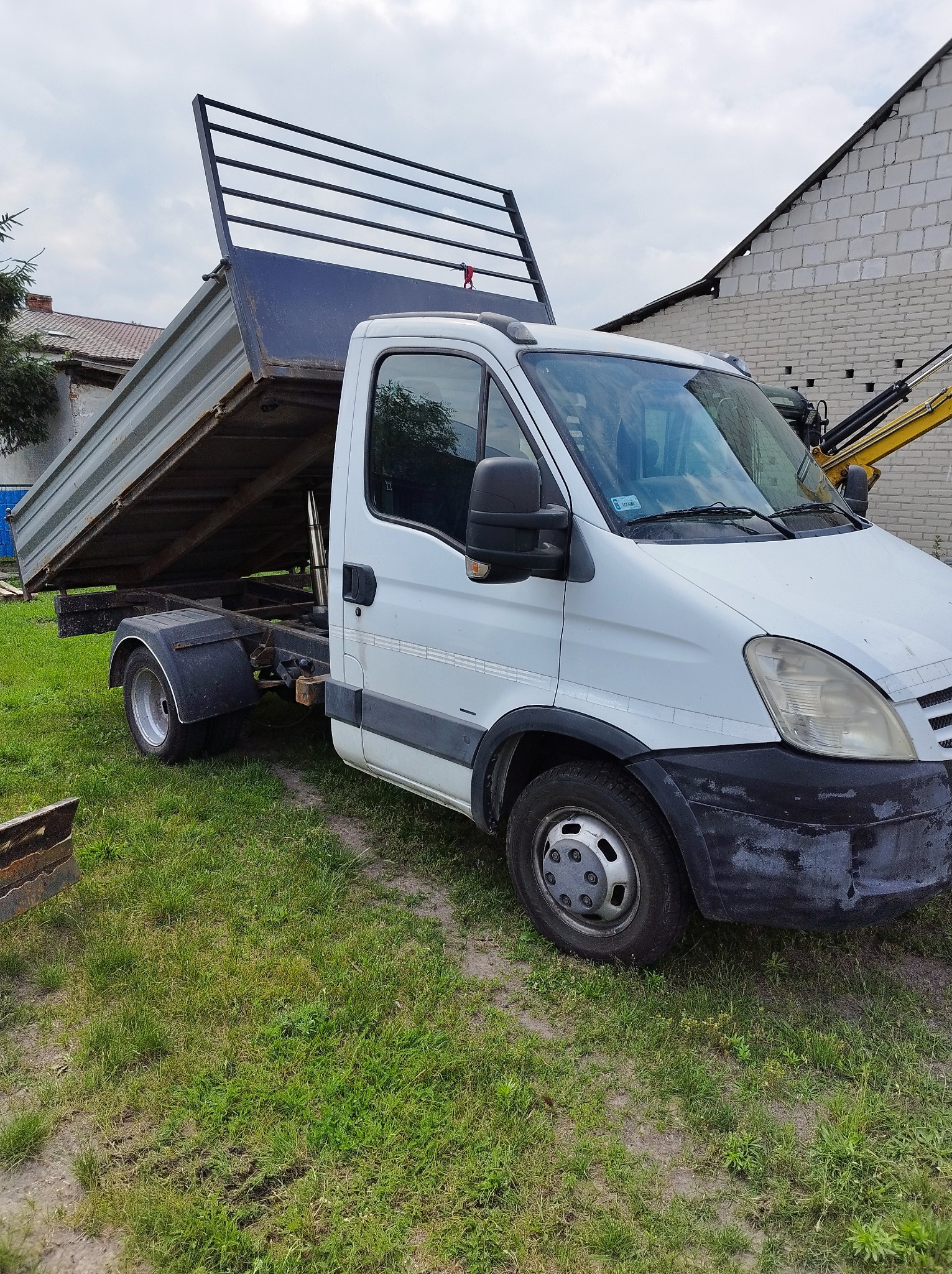 Biały samochód ciężarowy z podniesioną skrzynią ładunkową na trawiastym podłożu, w tle budynek z białej cegły i fragment koparki.