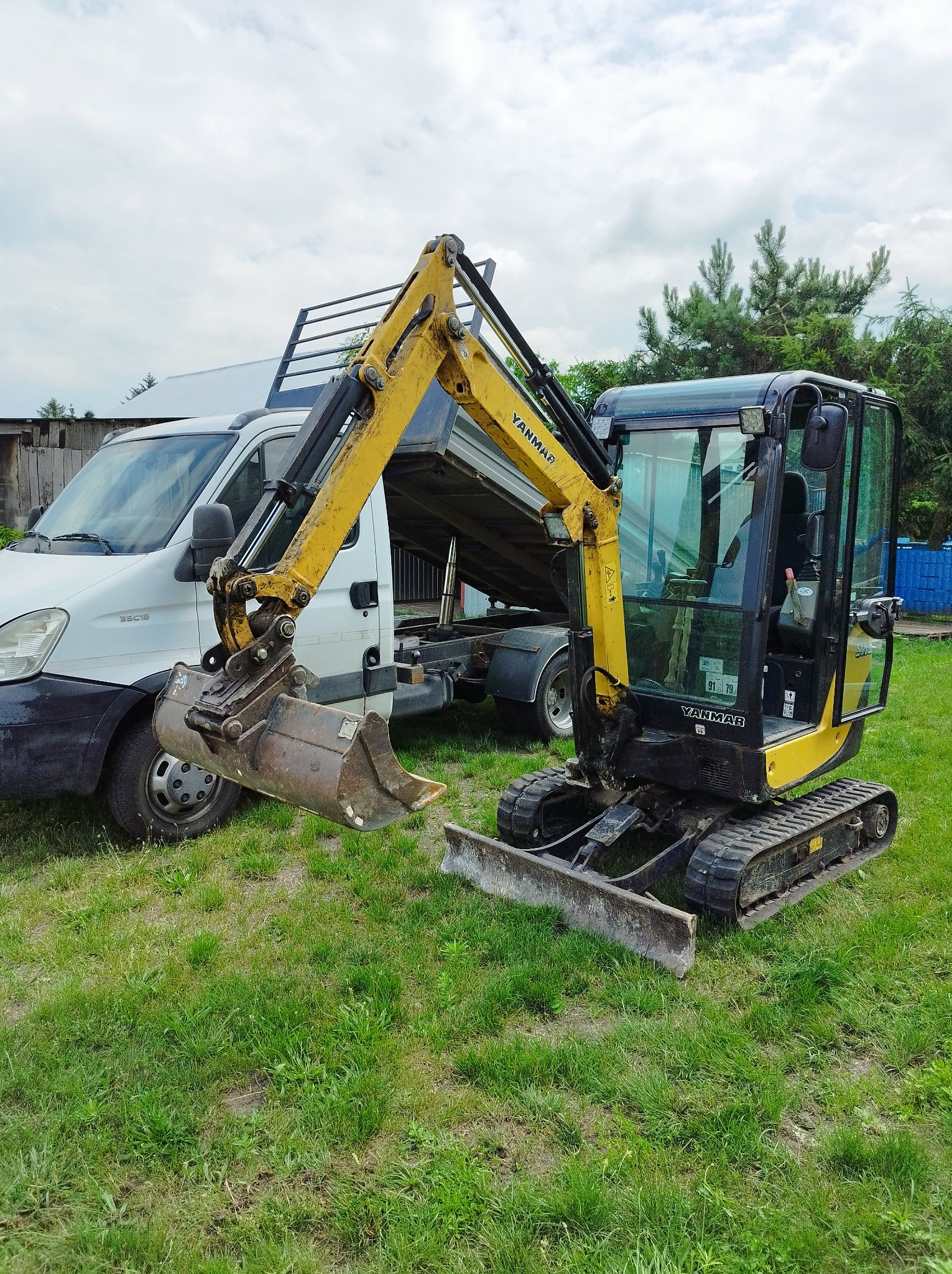 Żółto-czarna mini koparka Yanmar na trawie, z przodu lemiesz, z tyłu biały samochód dostawczy z otwartą skrzynią, pochmurne niebo.