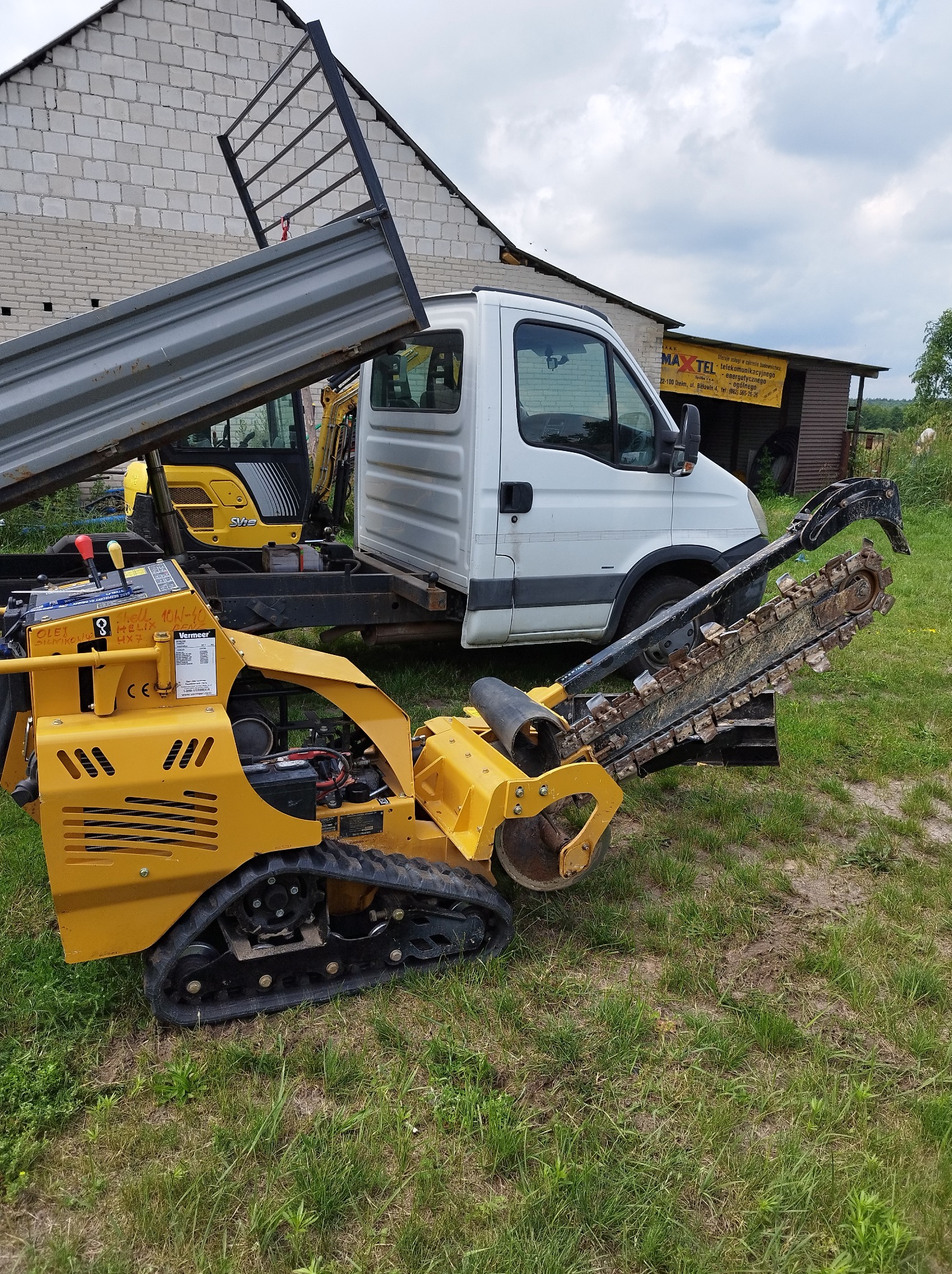 Żółta mini koparka łańcuchowa Vermeer na gąsienicach, zaparkowana obok białego samochodu dostawczego z otwartą skrzynią ładunkową, w tle budynek gospodarczy.