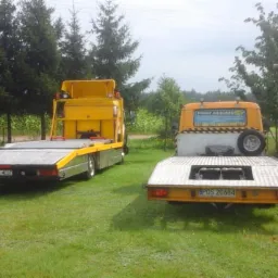 Dwie żółte lawety zaparkowane na trawie, jedna z reklamą 'Pomoc Drogowa' na kabinie, druga z widoczną tablicą rejestracyjną, w tle drzewa i pole kukurydzy.