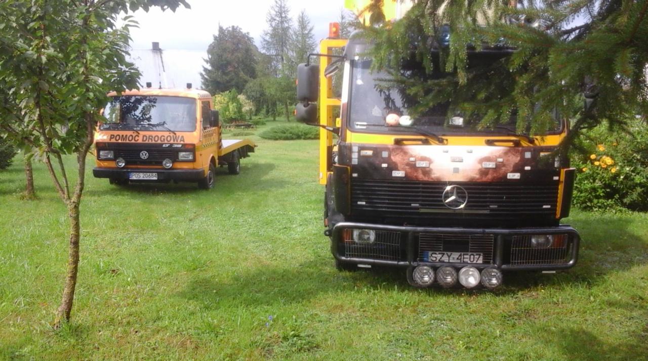 Dwie lawety na trawiastym terenie: żółta Volkswagen LT 35 z napisem 'Pomoc Drogowa' w tle i czarna Mercedes-Benz z przodu, częściowo zasłonięta przez gałęzie drzewa.