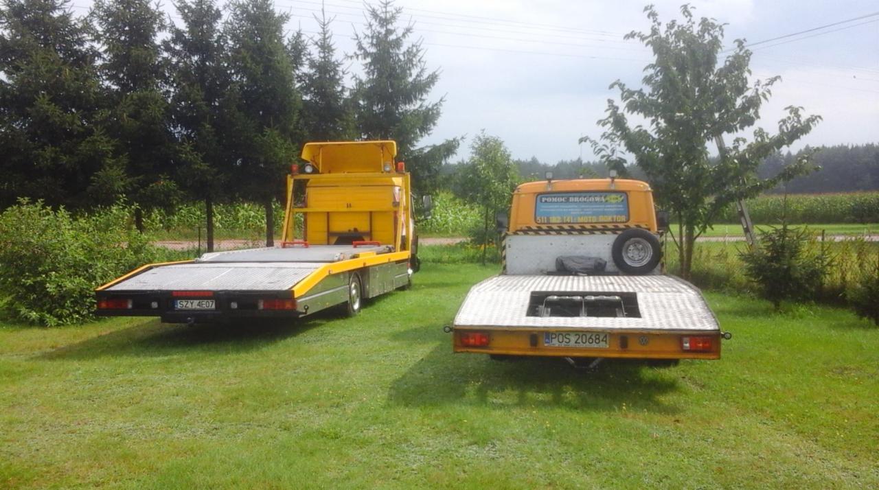 Dwie żółte lawety zaparkowane na trawie, jedna z reklamą 'Pomoc Drogowa' na kabinie, druga z widoczną tablicą rejestracyjną, w tle drzewa i pole kukurydzy.
