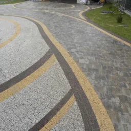 Ułożona nawierzchnia z kostki brukowej w odcieniach szarości i beżu, z dekoracyjnymi łukami i okręgami, prowadząca do posesji z widocznym fragmentem samochodu i trawnikiem.