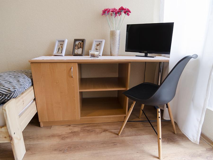 Pokój z łóżkiem, szafką, telewizorem i krzesłem w stylu skandynawskim, dekorowany ramkami ze zdjęciami i wazonem z kwiatami.