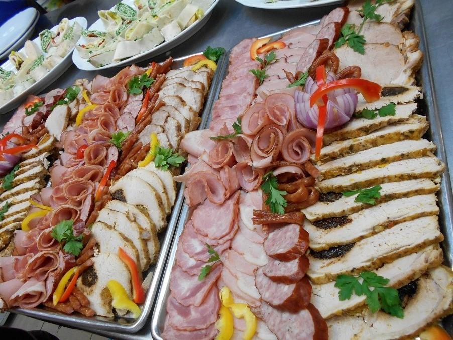 Dwie metalowe tace z różnorodnymi wędlinami, mięsami pieczonymi i roladkami, udekorowane papryką i natką pietruszki, na stole z białymi talerzami z roladkami w tle.