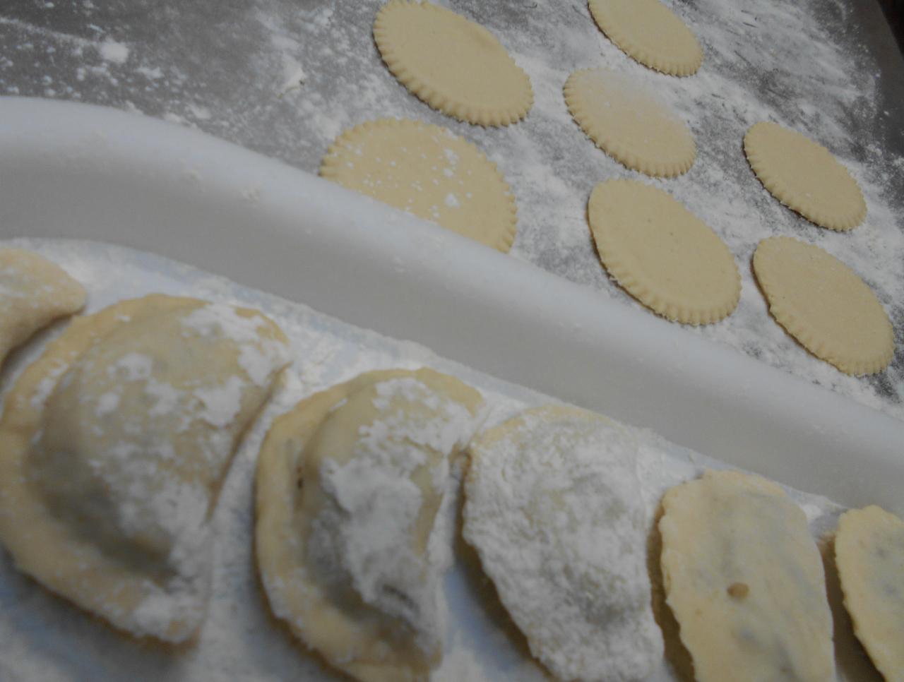 Przygotowywanie pierogów: surowe, okrągłe ciasto z karbowanymi brzegami obok uformowanych pierogów posypanych mąką na białej tacy.