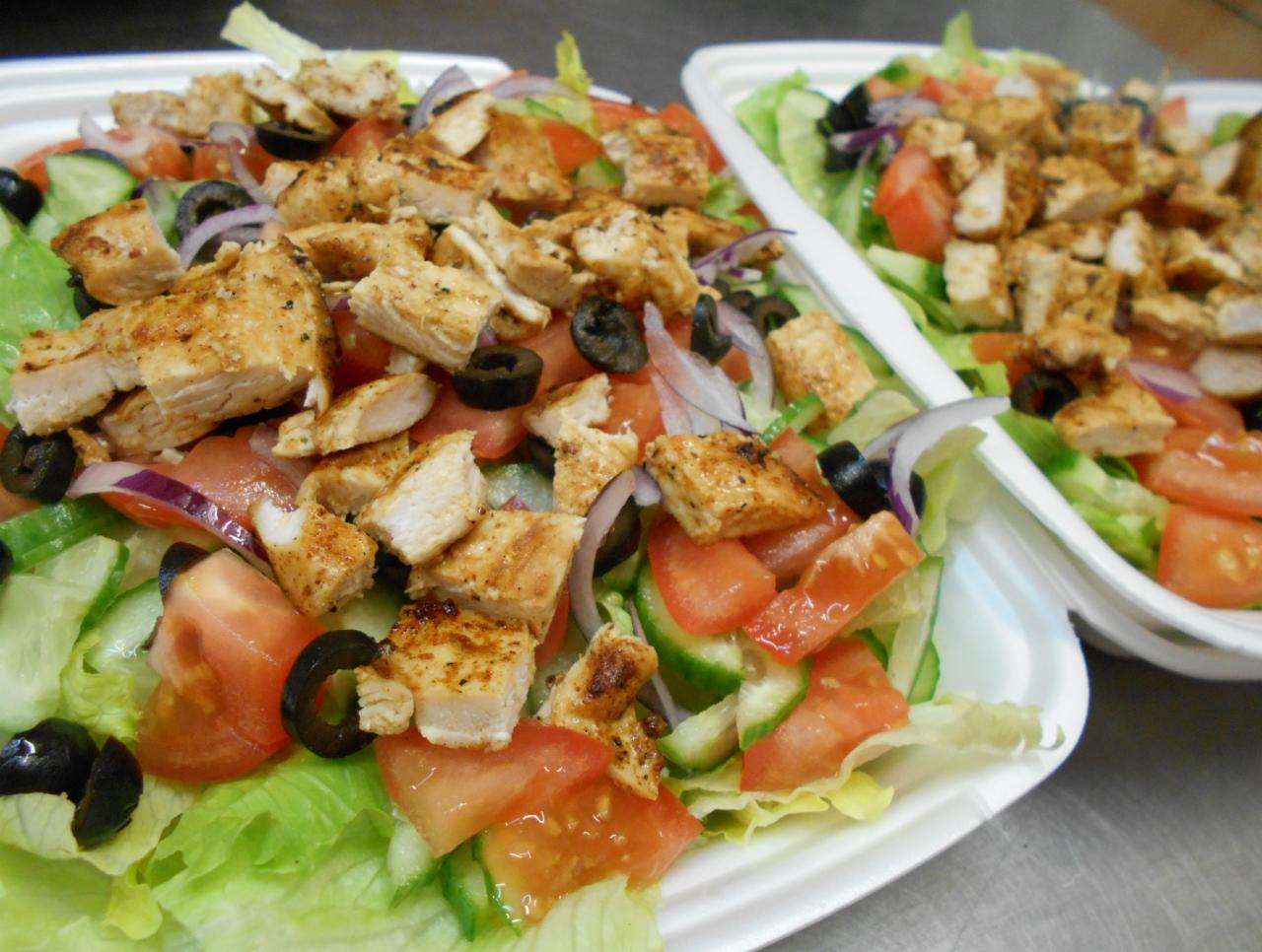 Dwie porcje sałatki w białych pojemnikach na wynos, z grillowanym kurczakiem w kostkach, pomidorami, ogórkami, czerwoną cebulą i oliwkami na podkładzie z sałaty lodowej.