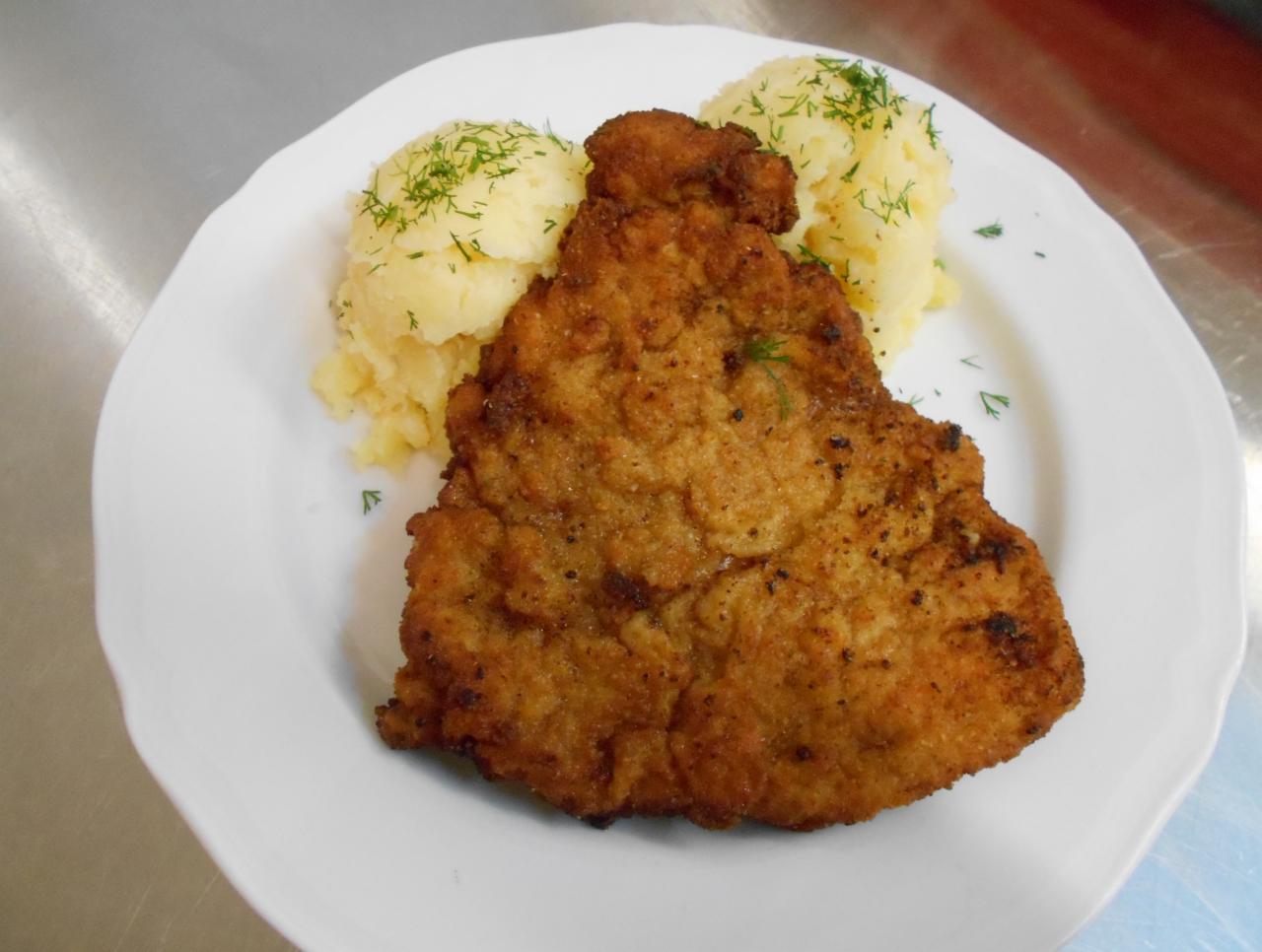 Duży, panierowany kotlet schabowy z dwiema porcjami ziemniaczanego puree, posypany koperkiem, podany na białym talerzu na metalowym blacie.