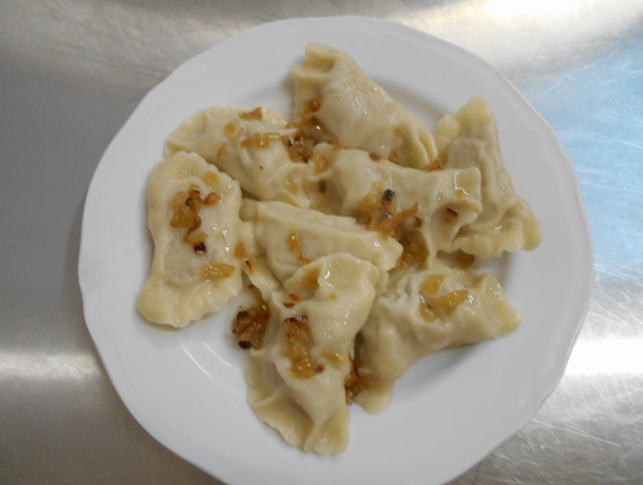 Porcja ręcznie robionych pierogów z farszem i podsmażoną cebulką na białym talerzu, widoczne delikatne zagniecenia ciasta.