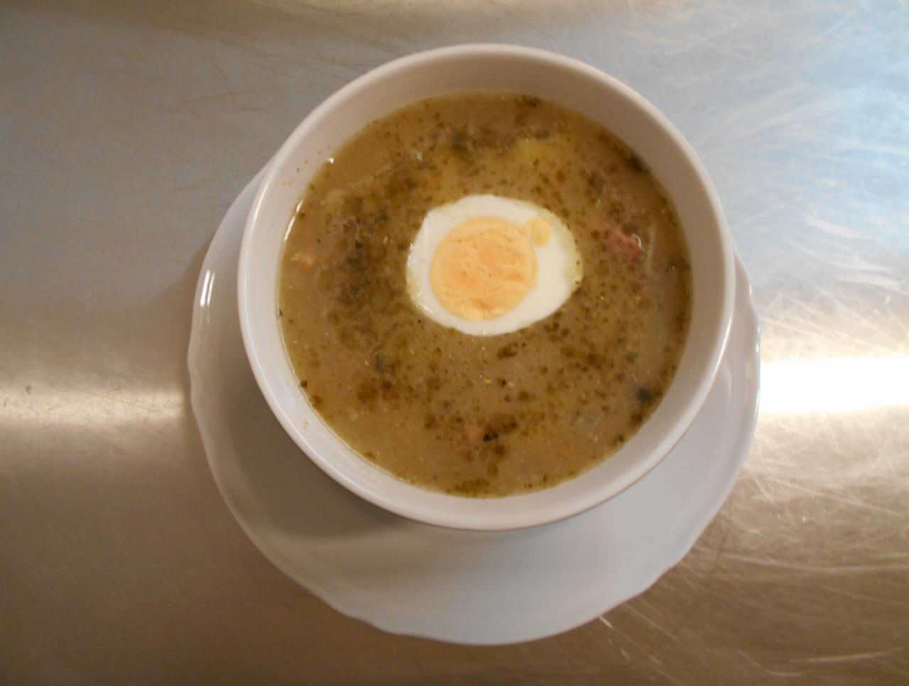 Żurek w białej miseczce na białym talerzu, z połówką jajka na twardo, na metalowym stole.