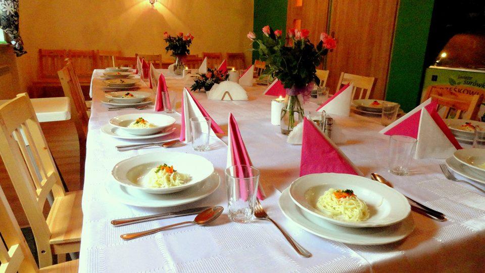 Uroczysty stół z białym obrusem, nakryty dla wielu gości, z talerzami z makaronem, różowymi serwetkami, szklankami i bukietami róż w wazonach.