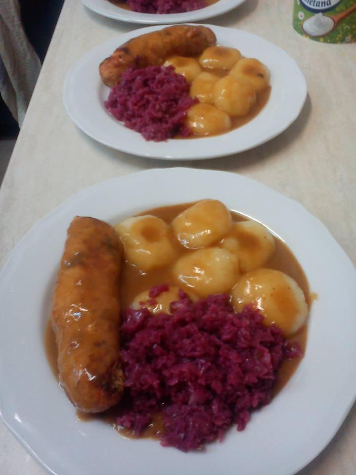 Dwa talerze z obiadem: długa kiełbasa w sosie, gotowane ziemniaki w sosie i czerwona kapusta, widok z góry, słabe oświetlenie.