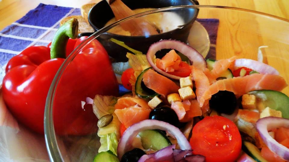 Szklana misa z sałatką z łososiem, czerwoną papryką, ogórkiem, czerwoną cebulą, pomidorami i grzankami, obok fondue serowe w czarnym garnku, na drewnianym stole z niebieską ściereczką w kratę.