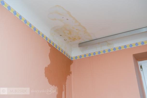 Zacieki na suficie i ścianie w rogu pokoju, widoczne plamy i uszkodzenia. Dekoracyjny pasek przy suficie. Problem z wilgocią w mieszkaniu.
