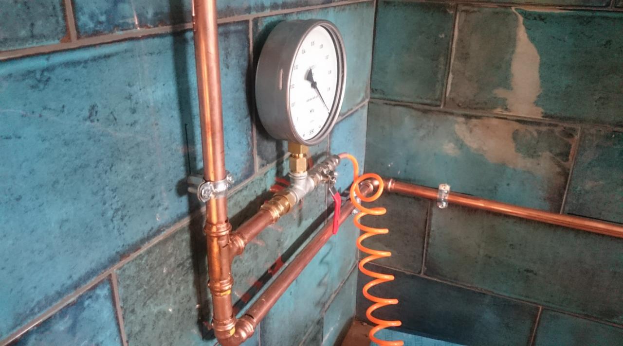 Miedziane rury wodne z manometrem na tle turkusowych płytek, instalacja hydrauliczna w łazience, kontrola szczelności instalacji wodnej po odbiorze mieszkania.