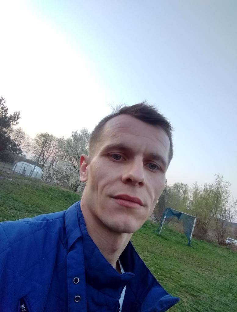 Portret mężczyzny w niebieskiej kurtce na tle zielonego trawnika i drzew. Ujęcie z dołu, widoczne niebo i fragment boiska.