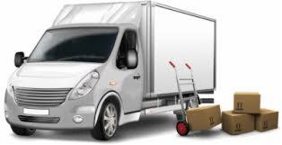 Szary samochód dostawczy z białą zabudową kontenerową, obok wózek transportowy z trzema kartonami, dodatkowe kartony na ziemi.