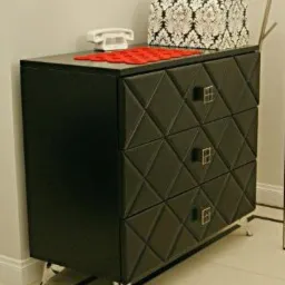 Czarna komoda z geometrycznym wzorem na frontach szuflad, metalowe uchwyty i nóżki, na blacie leży biały telefon retro i wzorzyste pudełko.