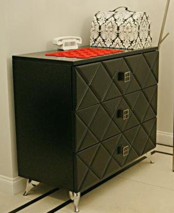 Czarna komoda z geometrycznym wzorem na frontach szuflad, metalowe uchwyty i nóżki, na blacie leży biały telefon retro i wzorzyste pudełko.