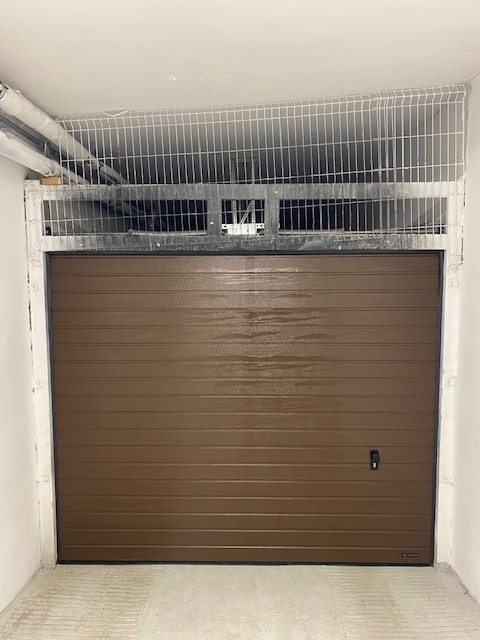 Zamknięta, brązowa brama garażowa segmentowa z czarnym uchwytem, widoczna krata zabezpieczająca nad bramą w garażu.