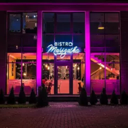 Fasada Bistro Marszałka oświetlona fioletowym neonem, widok na wnętrze z elementami dekoracyjnymi i schodami, nocne ujęcie z zewnątrz.