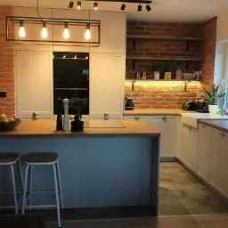 Nowoczesna kuchnia z ceglaną ścianą, drewnianym blatem i szarymi szafkami, oświetlona lampami wiszącymi i taśmą LED pod półkami.