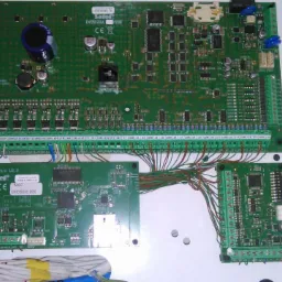 Otwarta obudowa centrali alarmowej z widocznymi płytkami drukowanymi, licznymi złączami i przewodami, wewnątrz biała obudowa.